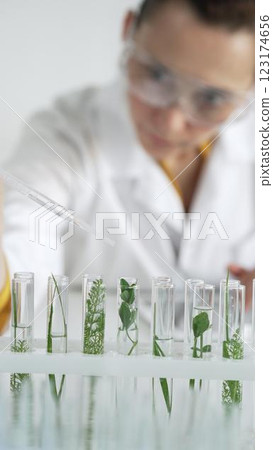 Woman scientist wearing a lab coat, is dripping liquid from a pipette into a test tube with a green plant inside, vertical close up view. Science and medicine concepts 123174656