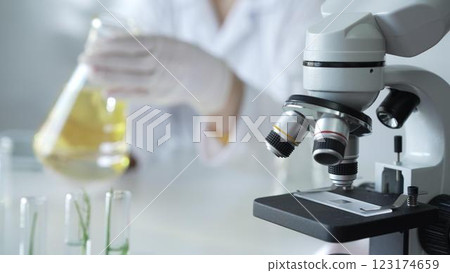 Microscope is near a scientist, wearing a lab coat and white protective gloves, and holding beaker with a yellow oily liquid in laboratory setting, close up. Medicine and science concept Microscope is near a scientist, wearing a lab coat and white protective gloves, and holding beaker with a yellow oily liquid in laboratory setting, close up. Medicine and science concept 123174659
