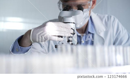 Man scientist researcher wearing white gloves, protective glasses and medical mask is using microscope in modern laboratory, portrait view. Low key lighting photo. Science and Medicine 123174666