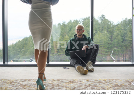 A blonde man sitting casually on the floor of a modern office, leaning against a window as he uses his laptop, with his attractive female colleague standing nearby 123175322