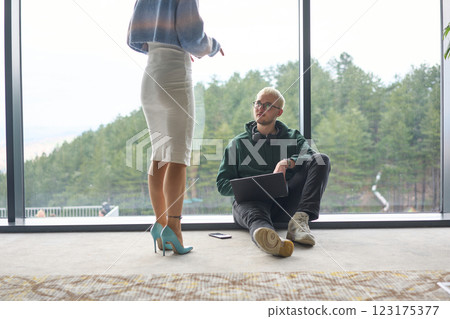 A blonde man sitting casually on the floor of a modern office, leaning against a window as he uses his laptop, with his attractive female colleague standing nearby A blonde man sitting casually on the floor of a modern office, leaning against a window as he uses his laptop, with his attractive female colleague standing nearby 123175377