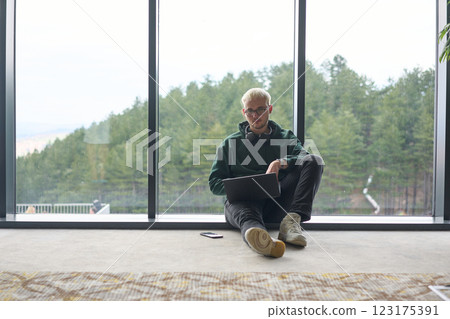 A blonde man sitting on the floor of a modern office, leaning against the window with a laptop in his lap, fully engaged in his work while maintaining a relaxed and focused demeanor. A blonde man sitting on the floor of a modern office, leaning against the window with a laptop in his lap, fully engaged in his work while maintaining a relaxed and focused demeanor. 123175391