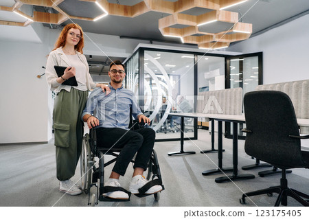 Business colleagues, collaborative business colleagues, including a person in a wheelchair, walk past a modern glass office corridor, illustrating diversity, teamwork and empowerment in the workplace. Business colleagues, collaborative business colleagues, including a person in a wheelchair, walk past a modern glass office corridor, illustrating diversity, teamwork and empowerment in the workplace. 123175405