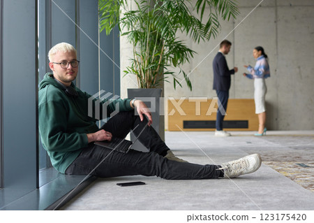A casual young professional sitting by a large window in a modern office, smiling while working on a laptop, with colleagues interacting in the blurred background. 123175420