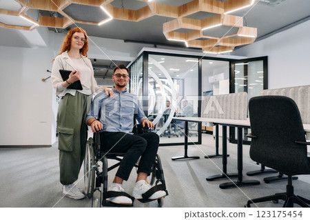 Business colleagues, collaborative business colleagues, including a person in a wheelchair, walk past a modern glass office corridor, illustrating diversity, teamwork and empowerment in the workplace. 123175454