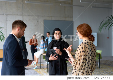 A diverse group of business professionals engages in a conversation during a work break in the office, with their colleagues visible in the background, fostering collaboration and connection 123175455