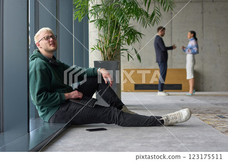 A casual young professional sitting by a large window in a modern office, smiling while working on a laptop, with colleagues interacting in the blurred background. A casual young professional sitting by a large window in a modern office, smiling while working on a laptop, with colleagues interacting in the blurred background. 123175511