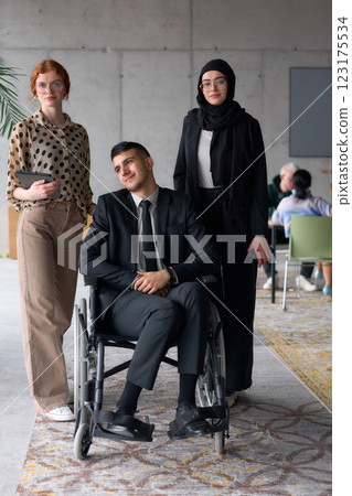 A diverse business team poses confidently, including a smiling director seated in a wheelchair, flanked by two professional female colleagues, one wearing a hijab and the other holding a tablet, in a 123175534