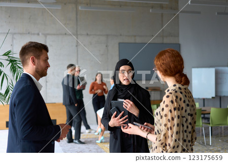 A diverse group of business professionals engages in a conversation during a work break in the office, with their colleagues visible in the background, fostering collaboration and connection 123175659