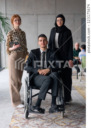 A diverse business team poses confidently, including a smiling director seated in a wheelchair, flanked by two professional female colleagues, one wearing a hijab and the other holding a tablet, in a 123175674