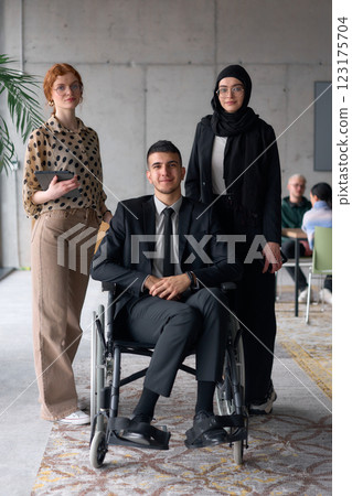 A diverse business team poses confidently, including a smiling director seated in a wheelchair, flanked by two professional female colleagues, one wearing a hijab and the other holding a tablet, in a A diverse business team poses confidently, including a smiling director seated in a wheelchair, flanked by two professional female colleagues, one wearing a hijab and the other holding a tablet, in a 123175704