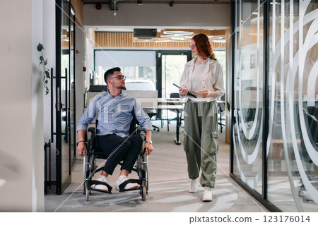 Business colleagues, collaborative business colleagues, including a person in a wheelchair, walk past a modern glass office corridor, illustrating diversity, teamwork and empowerment in the workplace. 123176014
