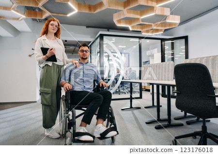 Business colleagues, collaborative business colleagues, including a person in a wheelchair, walk past a modern glass office corridor, illustrating diversity, teamwork and empowerment in the workplace. Business colleagues, collaborative business colleagues, including a person in a wheelchair, walk past a modern glass office corridor, illustrating diversity, teamwork and empowerment in the workplace. 123176018