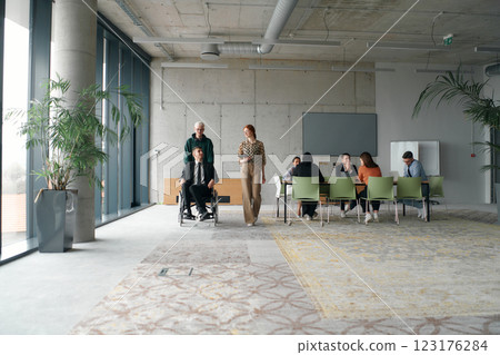 A diverse and multicultural business team collaborates in the office while their colleagues, including a director in a wheelchair and his friend, share a lighthearted moment, fostering an atmosphere 123176284