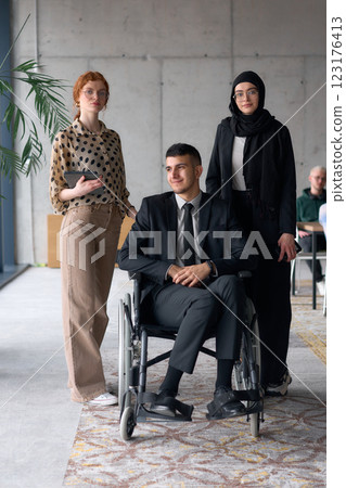 A diverse business team poses confidently, including a smiling director seated in a wheelchair, flanked by two professional female colleagues, one wearing a hijab and the other holding a tablet, in a 123176413