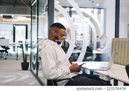 African American businessman wearing headphones while using a smartphone, fully engaged in his work at a modern office, showcasing focus, productivity, and contemporary professionalism African American businessman wearing headphones while using a smartphone, fully engaged in his work at a modern office, showcasing focus, productivity, and contemporary professionalism 123176434