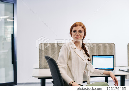 A modern businesswoman with striking orange hair sitting confidently in a contemporary office, accompanied by her laptop, embodying professionalism, creativity, and success in a technology-driven 123176465