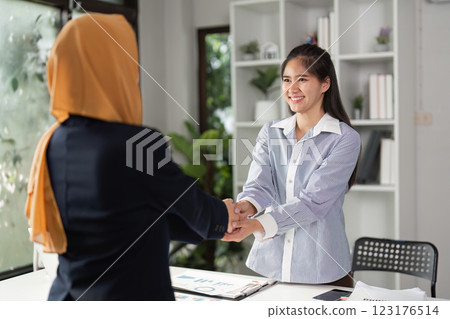 Diverse businesswomen smiling while shaking hands in a modern office, highlighting collaboration and partnership. 123176514