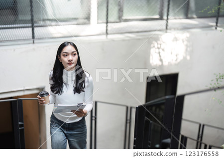 Young professional woman walking up stairs with documents and coffee cup, focused on her work and planning. Young professional woman walking up stairs with documents and coffee cup, focused on her work and planning. 123176558