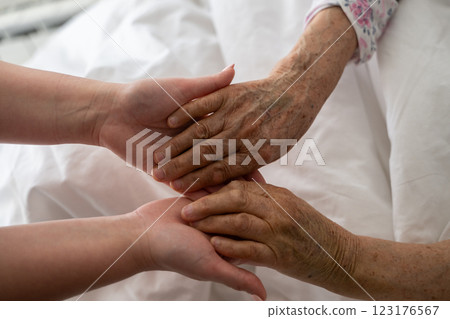 A young womans hands gently hold the hands of an elderly female patient in a hospital bed, symbolizing care, support, and compassion. 123176567