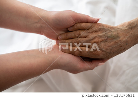 A young womans hands gently hold the hands of an elderly female patient in a hospital bed, symbolizing care, support, and compassion. 123176631
