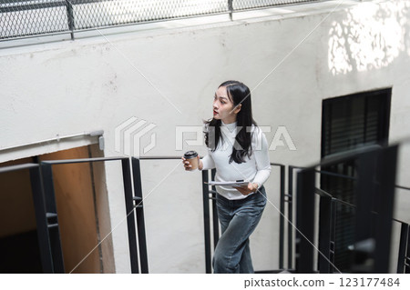 Young Asian woman ascending spiral staircase with coffee and documents, reflecting on work and daily life. Young Asian woman ascending spiral staircase with coffee and documents, reflecting on work and daily life. 123177484