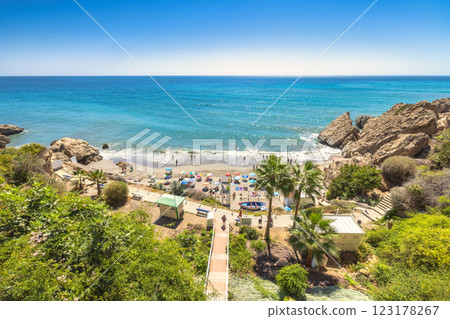 Nerja town, a resort of The Costa del Sol seaside region in Andalusia in the south of Spain. Secluded cove beach with turquoise water, surrounded by cliffs and palm trees. People relax on sandy shore. 123178267