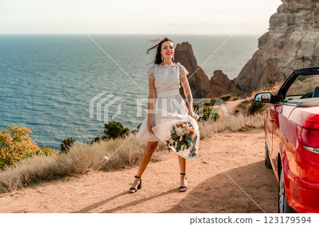 A woman in a white dress is standing next to a red convertible car. She is holding a bouquet of flowers and she is in a happy mood. The scene suggests a romantic or special occasion. A woman in a white dress is standing next to a red convertible car. She is holding a bouquet of flowers and she is in a happy mood. The scene suggests a romantic or special occasion. 123179594