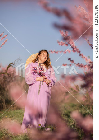 Woman blooming peach orchard. Against the backdrop of a picturesque peach orchard, a woman in a long pink dress and hat enjoys a peaceful walk in the park, surrounded by the beauty of nature. 123179596
