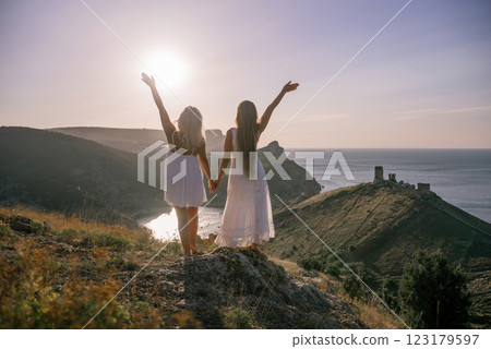 Two women are standing on a hill overlooking the ocean. They are holding hands and looking out at the water. The scene is peaceful and serene, with the sun shining brightly in the background. Two women are standing on a hill overlooking the ocean. They are holding hands and looking out at the water. The scene is peaceful and serene, with the sun shining brightly in the background. 123179597