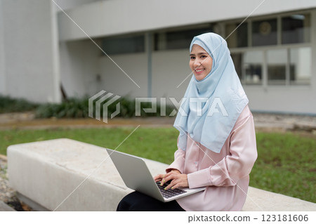 Muslim woman happily working on her laptop outdoors, promoting a positive work environment. Muslim woman happily working on her laptop outdoors, promoting a positive work environment. 123181606