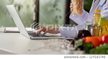 Nutritionist typing on laptop while holding whole grain pasta, focusing on healthy meal planning and dietary advice. 123181746