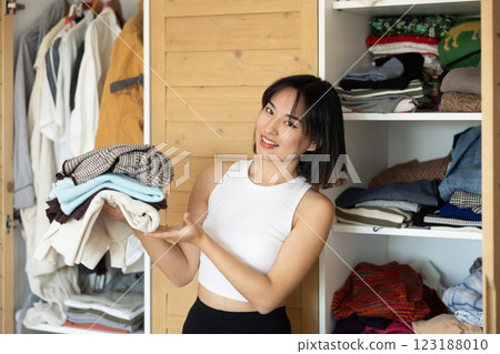 Happy young Asian girl holding some items of clothing by the wardrobe Happy young Asian girl holding some items of clothing by the wardrobe 123188010