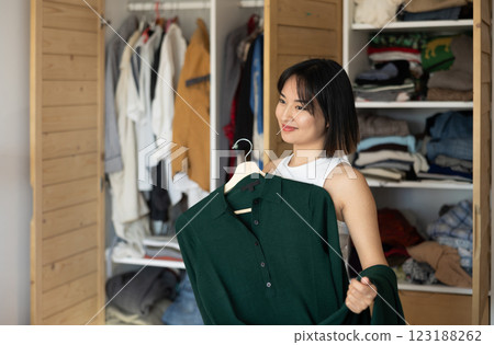 Happy young Asian girl trying to choose a dress standing by wardrobe 123188262