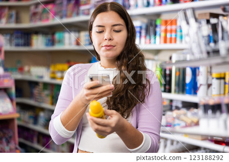 Pretty young woman looking for lotion in cosmetics store and taking photo on mobile phone Pretty young woman looking for lotion in cosmetics store and taking photo on mobile phone 123188292