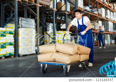 In warehouse of store, stevedore loader young man pushes and carries large cart for bulky cargo In warehouse of store, stevedore loader young man pushes and carries large cart for bulky cargo 123188337