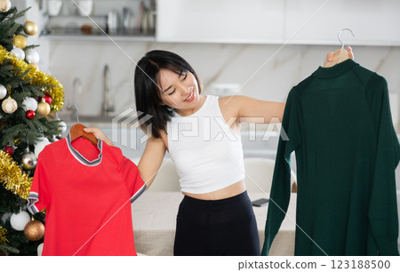 Happy young Asian girl trying to choose a dress standing next to Xmas-tree 123188500