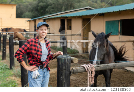 Female stable worker stands near wooden fence of paddock and watches horse. Female stable worker stands near wooden fence of paddock and watches horse. 123188503