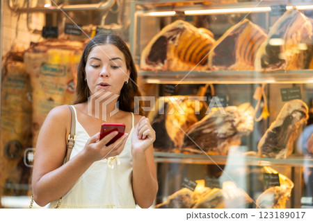 Girl with phone in hands looks at shop window, examines dried beef, looks at screen of smartphone Girl with phone in hands looks at shop window, examines dried beef, looks at screen of smartphone 123189017