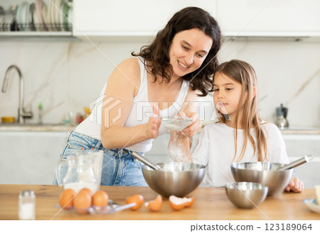 Happy mother and her daughter adding flour in metal bowl and whisking ingredients in the kitchen Happy mother and her daughter adding flour in metal bowl and whisking ingredients in the kitchen 123189064