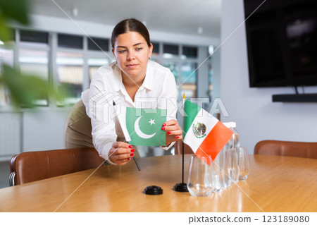 A woman in a white blouse with friendly smile arrangs flags of Mexico and Pakistan for meeting. 123189080
