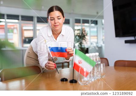 A woman in a white blouse with friendly smile arrangs flags of Russia and Iran for meeting. 123189092