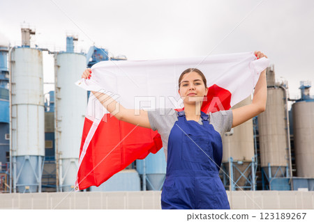 near large plant in Polish province,calm girl in blue overalls stands and holds flag of Poland 123189267