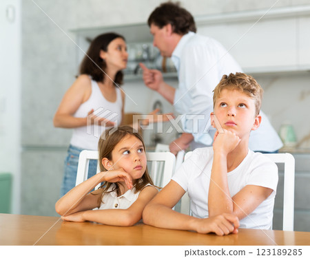 Two children sitting at the kitchen table sadly while their parents quarrelling Two children sitting at the kitchen table sadly while their parents quarrelling 123189285