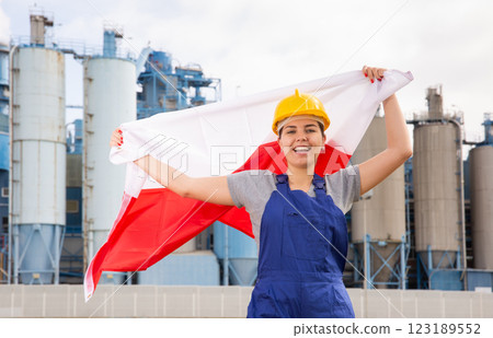 Young female engineer in helmet waving state flag of Poland while standing in front of big tanks at chemical plant 123189552