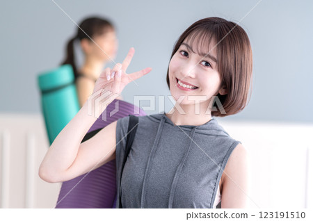 年輕女子微笑著，手拿瑜珈墊，做出 V 形手勢 123191510