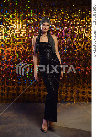 Elegant woman poses in shimmering black dress against a colorful sequin backdrop at a fashion event 123192600
