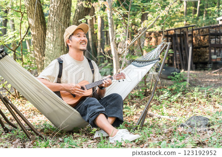 亞洲男子坐在露營地/戶外/休閒的吊床上彈奏尤克里裡琴 123192952
