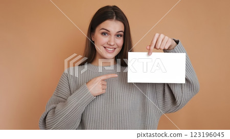Cheerful Young Woman in Knitted Sweater Pointing at Blank Card Against Solid Background - Mockup Template for Advertisement or Announcement 123196045