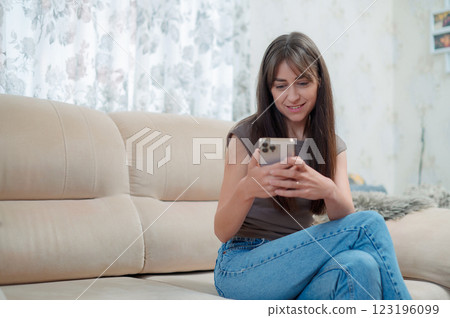 Young happy woman sitting comfortably on sofa, smiling while using her smart phone indoors during a relaxing afternoon Young happy woman sitting comfortably on sofa, smiling while using her smart phone indoors during a relaxing afternoon 123196099